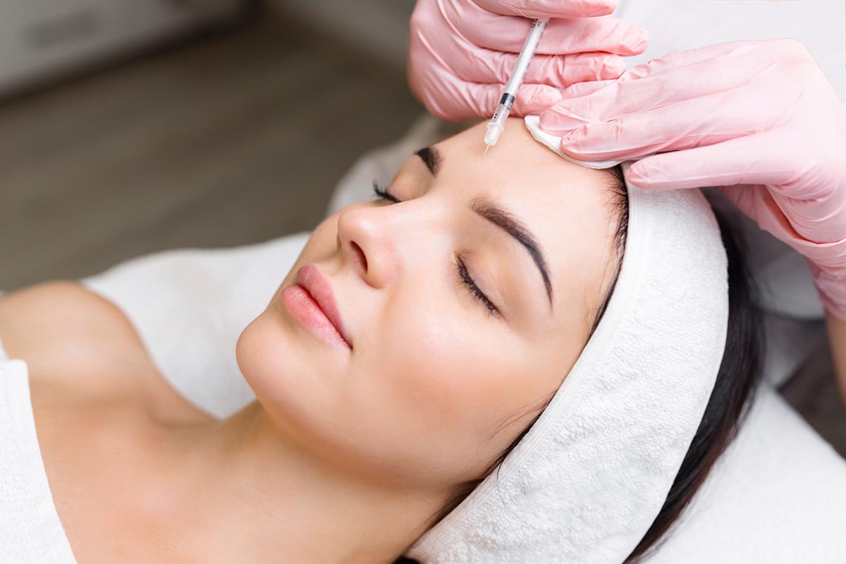 Close up of hands of young cosmetologist injecting botox in female face. She is standing and smiling. The woman is closed her eyes with relaxation.