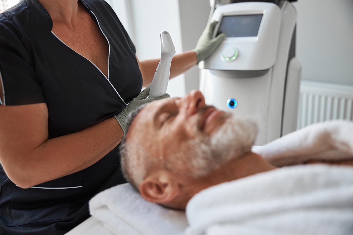 Professional beautician starting a machine for photofacial treatment while providing skincare for an elderly man